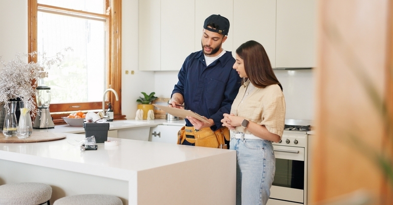 A service technician wearing a tool belt discusses paperwork with a woman in a bright kitchen.