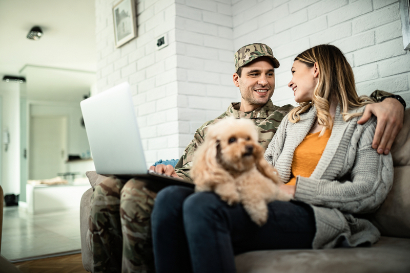 A smiling soldier in a uniform and a woman sit on a sofa with a small dog, looking at each other beside an open laptop.