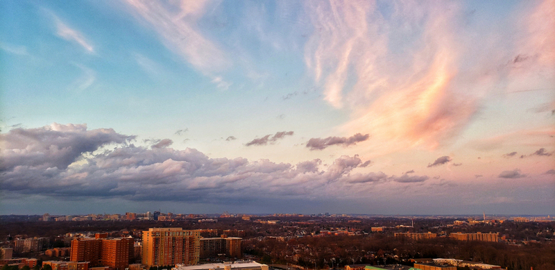 An aerial sunset view of a sprawling city skyline under a wide sky filled with dramatic pink, white, and dark grey clouds.