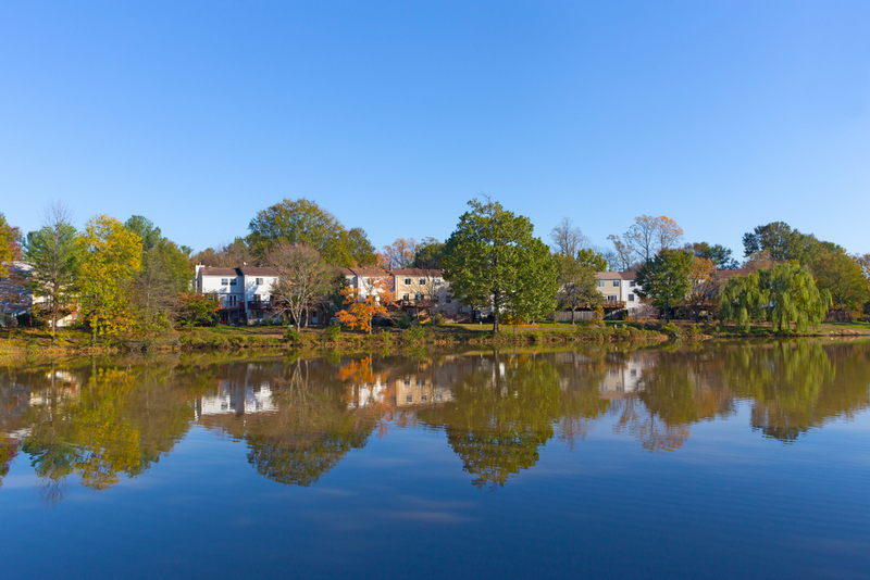 A row of suburban houses and colorful autumn trees reflected on the calm surface of a wide lake under a clear blue sky.