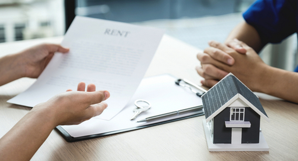 A real estate agent reviews a paper contract labeled "RENT" with a client at a desk featuring a small model house and keys.