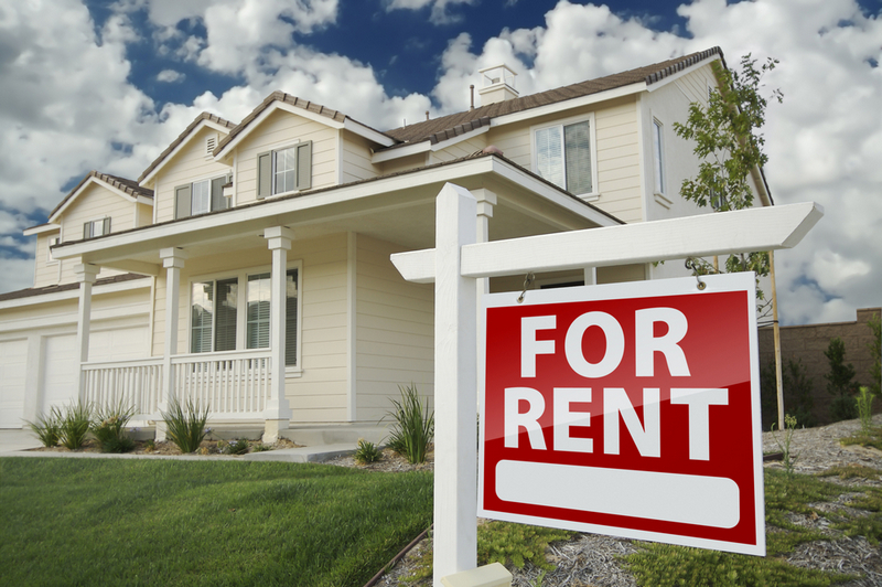 A red "For Rent" sign stands on a green lawn in front of a modern, two-story suburban home.