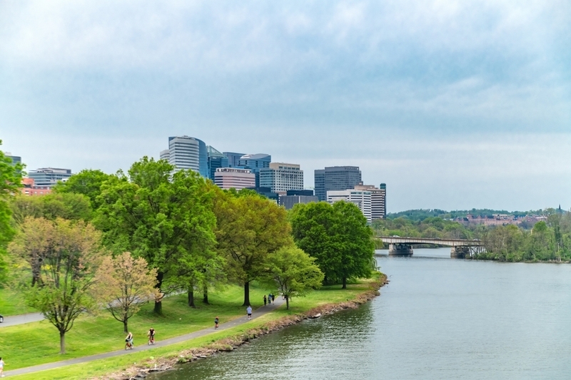 A wide shot of the Rosslyn skyline in Arlington, Virginia, viewed from across the Potomac River with a lush green park and walking path in the foreground under an overcast sky.