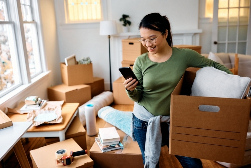 A smiling woman holding a moving box while looking at her smartphone in a room filled with cardboard boxes.
