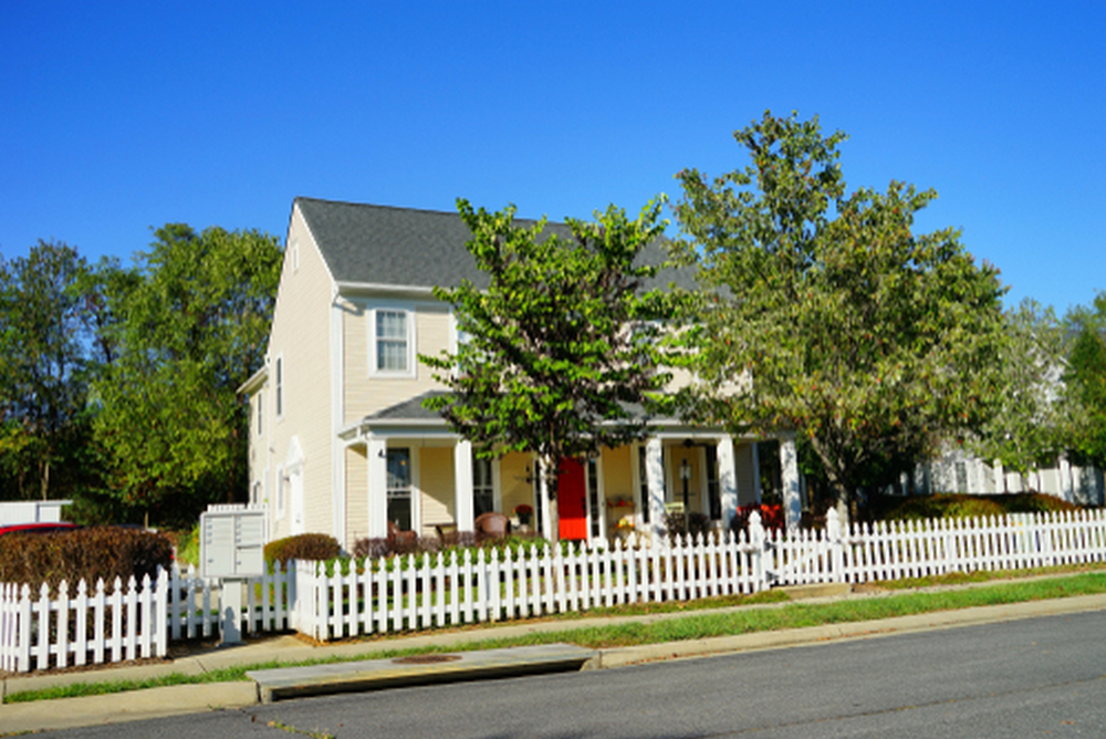 A two-story cream-colored house with a red front door, columned porch, and white picket fence under a clear blue sky.