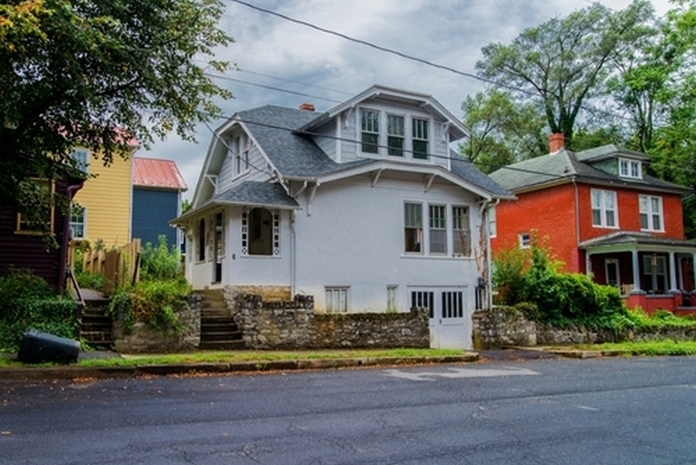 A street view of historic two-story houses in a residential neighborhood, featuring a central white home with a built-in garage next to a bright red house, set behind a stone retaining wall under a cloudy sky.