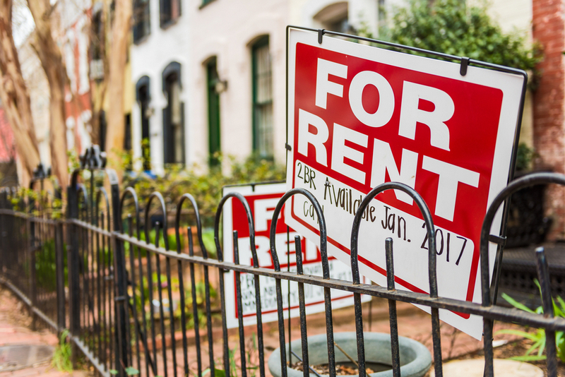 A red "FOR RENT" sign is displayed behind a black wrought-iron fence in front of urban row houses.If you plan to use this for a specific platform, let me