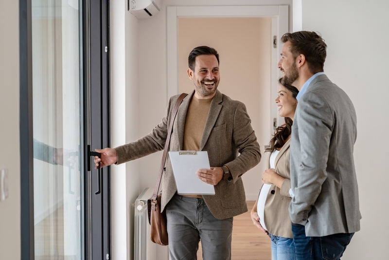 A smiling real estate agent opens a glass door to show a modern apartment to a young couple.