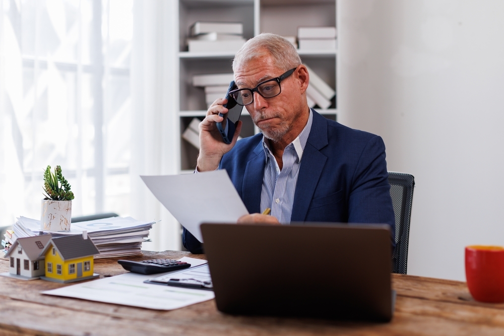 An older man in a blue suit and glasses looks at paperwork while talking on a smartphone at a desk with a laptop and miniature house models.