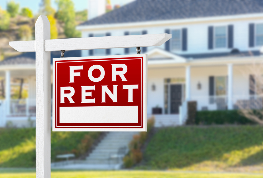 A red and white "For Rent" sign hanging from a white wooden post on a green lawn, with a large, two-story house blurred in the background.