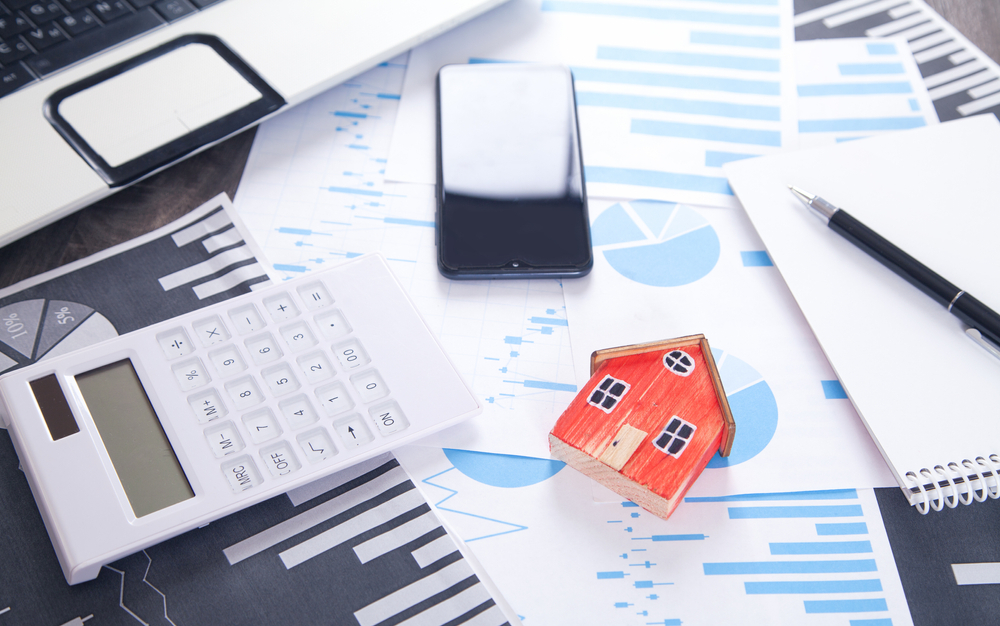 A white calculator, a tiny orange model house, a smartphone, a pen, and a notepad sit on top of sheets of paper printed with financial charts and graphs next to a laptop computer.