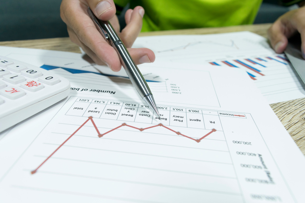 A close-up of a person's hand holding a pen and pointing at a printed line graph on a desk alongside a calculator and other financial charts.