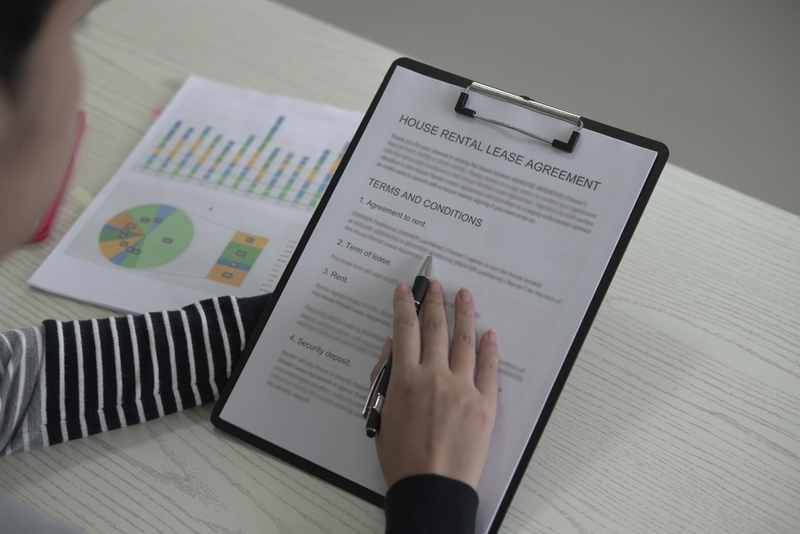 An overhead, close-up shot of a person's hand holding a pen and reviewing a "House Rental Lease Agreement" attached to a black clipboard on a wooden desk.