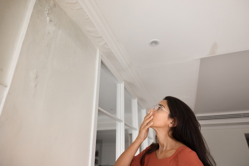 A concerned woman looking up at water damage and cracks on her apartment wall and ceiling.