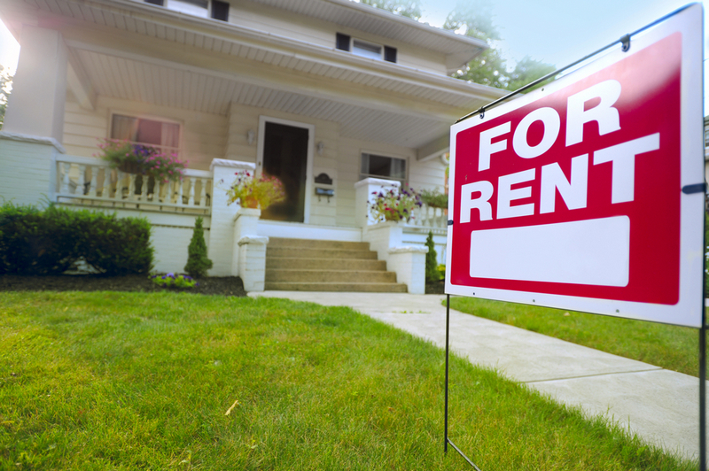 A red and white "FOR RENT" sign stands in the foreground on a green lawn next to a concrete walkway leading up to a two-story house with a front porch.