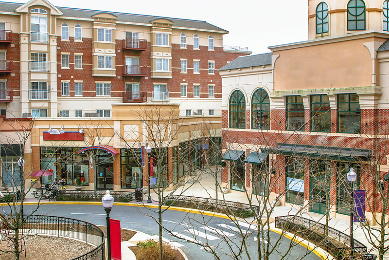 A street-level shot captures a modern mixed-use development featuring retail storefronts on the ground level and multi-story brick apartment buildings above, organized around a curved pedestrian plaza and roadway.