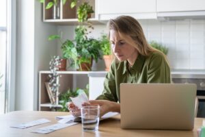 Thoughtful upset woman checking receipts and statements at kitchen table with water glass