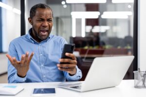 Stressed landlord looking at a phone, frustrated with rental issues or tenant problems while working at a laptop