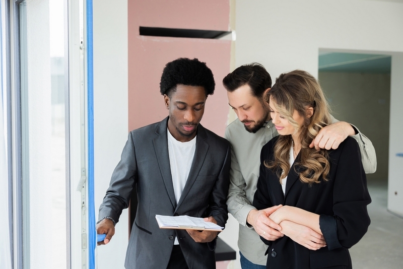 An African American real estate agent in a grey blazer shows documents to a couple inside an unfinished house.