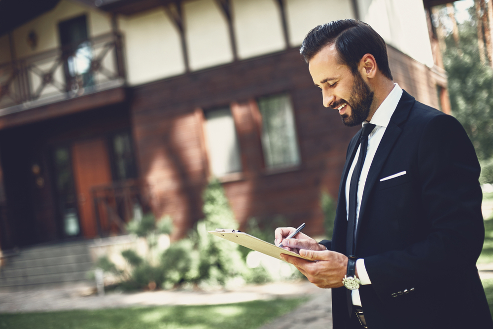 A smiling, bearded real estate agent in a black suit writes on a clipboard outdoors in front of a large house.