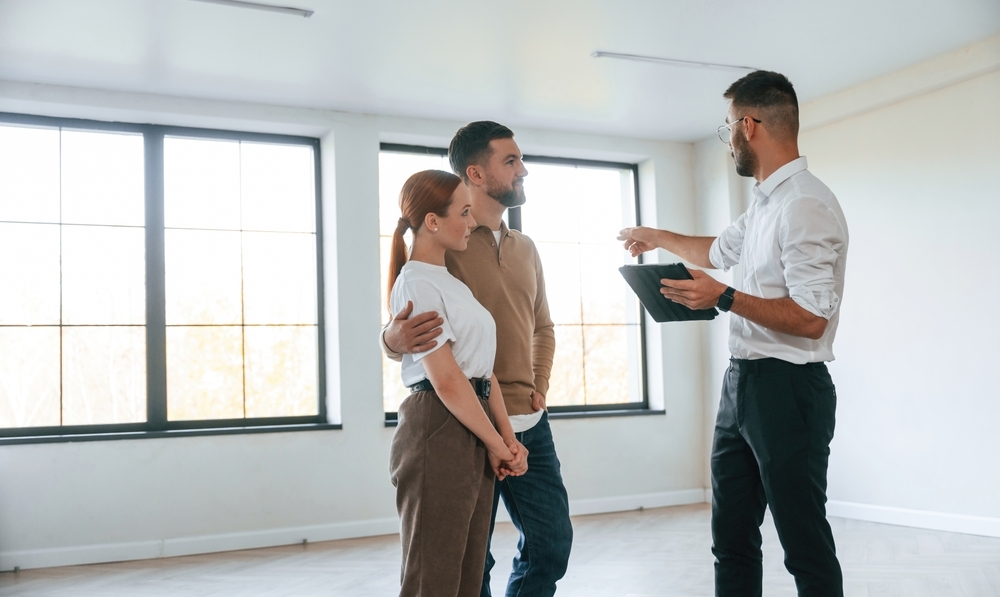 A real estate agent with a tablet shows an empty, sunlit apartment to a young couple.